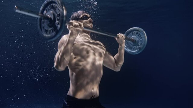 Cinematic shot of young male professional bodybuilder athlete with muscular body is lifting with effort and determination barbell underwater. Concept of sport, fitness, healthy lifestyle, recreation.
