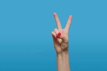 Female hand shows victory sign, letter V. Female hand with red gel polish manicure on a blue background. Copy space