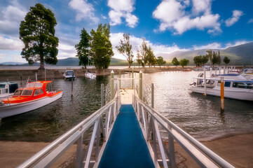Fototapeta na wymiar Jetty perspective and scenic view with backlit trees in the background at Te Anau Boating Club Marina, on the shore of Lake Te Anau, New Zealand, South Island, on a beautiful afternoon at golden hour.