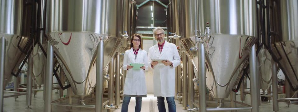 Two brewery technologists in a brewery wearing white lab coats. Brewery technologists looking at camera and smiling. Brewery, factory concept.