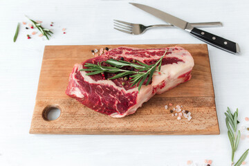 Raw ribeye steak on board, knife, fork, pepper, rosemary and pink Himalayan salt