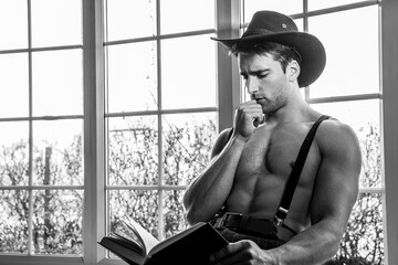 Portrait of handsome shirtless cowboy wearing hat sitting in window with book in lap, reading