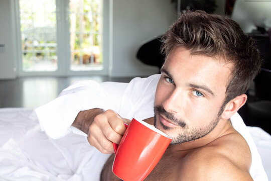Handsome hairy naked muscular man with beard sixpack abs lying in bed covered with sheet drinking coffee