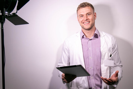 Portrait Of Handsome Male Doctor, Smiling And Looking At Camera With Behind The Scenes Studio Lamp In Shot