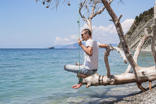 Summer landscape with a view of the sea. A guy sitting on a swing, on a fallen tree on the shore, and cargo ships on the horizon. The outskirts of the resort of Gelendzhik. Russia, Black Sea coast