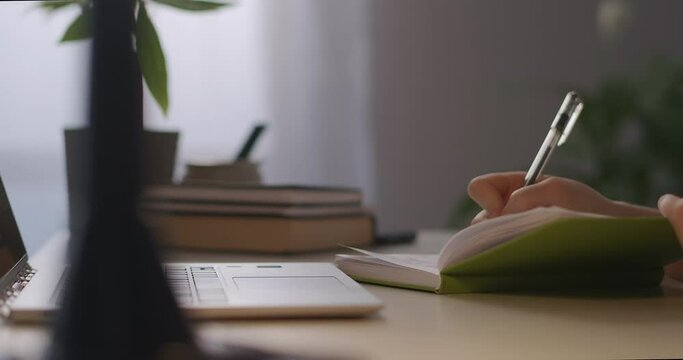 woman student is writing notes in notebook by pen, closeup of working table with laptop, e-learning and online education