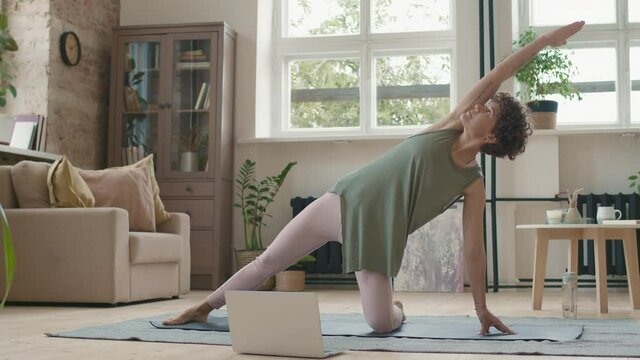 Tracking shot of fit young woman in sportswear watching online yoga class and doing gate pose on mat at home