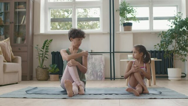 Tracking shot of fit young mother with short hair sitting on mat and talking to cute little daughter while practicing yoga with her in morning