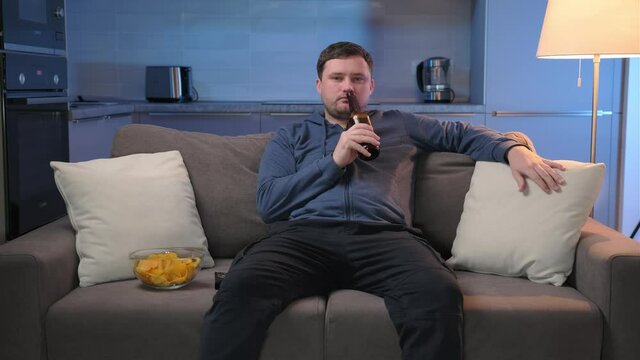 Man drinks beer and eats chips while sitting at home on couch, in evening he watches movies at home on TV. Unshaven caucasian man rests at home and watches TV shows or sports news on TV screen.