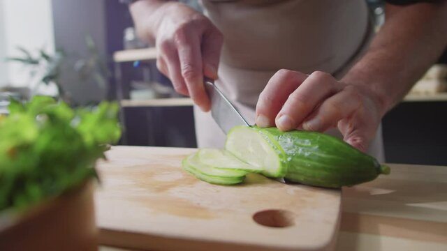 Close up shot of hands of male chef cutting fresh cucumber into thin slices with knife on wooden board in kitchen