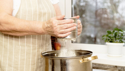 The concept of cooking at home. The hands of an elderly woman sift the flour for the dough.