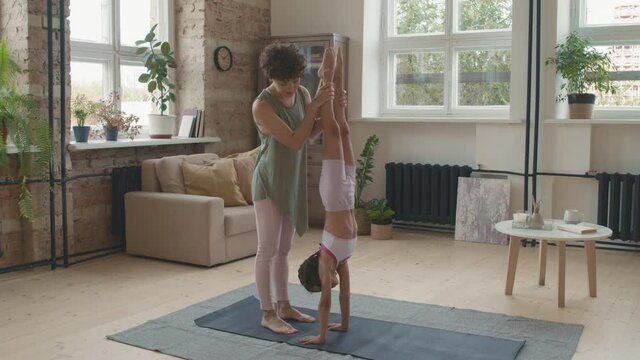 Slowmo medium shot of cheerful young mother with short curly hair helping her little daughter practicing handstand on yoga mat at home, then high-fiving her