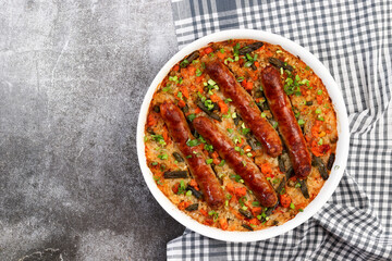 Oven baked sausages with rice and vegetables in a white baking dish on a dark grey background. Top view, flat lay.