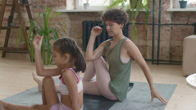 Medium shot of fit young mother and little daughter in sportswear sitting on yoga mat and doing half spinal twist pose while stretching together
