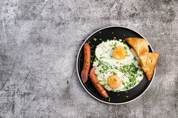 Fried eggs with green onions, sausages and toast on a round plate on a dark grey background. Top view, flat lay