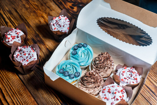 Delivery in carton box container of cupcakes or fairy cakes decorated with blueberries and blue creamy-cheese, brown cocoa topping and white cream on dark wooden table. Horizontal orientation image