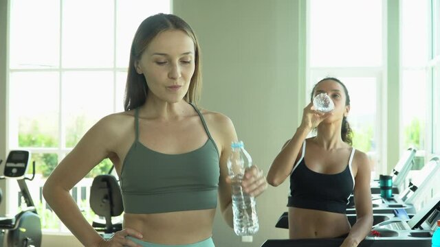 group of two young sports woman taking a break from running drinking water on a treadmill in fitness gym indoors. runner girl in sportswear rest after workout standing on running machine