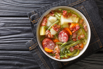 Homemade soup of green beans, potatoes, leeks, carrots with sausages and bacon close-up on a plate on the table. Horizontal top view from above