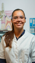 Stomatologist doctor smiling at camera in dental office while assistant preparing man for stomatological surgery in background. Woman dentist looking on webcam sitting on chair in dentist room