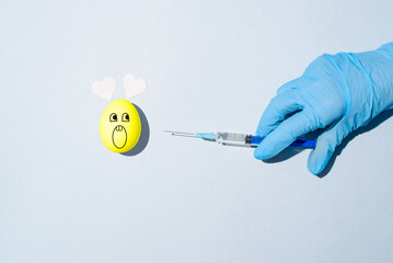 Easter quarantine concept. Funny egg with painted bunny face and heart shaped ears. Hand in medical glove holding syringe of vaccine. Injection.