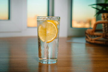 A glass of water with ice and lemon on a wooden table in front of the window with a beautiful light