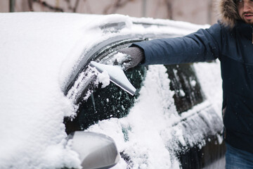 Close up of man is cleaning snowy window on a car with snow scraper. Focus on the scraper. Cold snowy and frosty morning. Black car