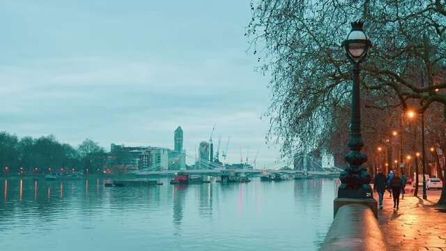 Timelapse over the River Thames and Albert Bridge in Battersea, London in 4K. A bridge being lighted and people walking on the sidewalk of a road in the evening.