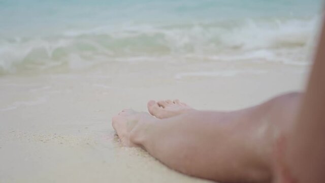Close up tan skin female legs sit dow on white sand beach, crystal clear sea water, relaxing summer tropical paradise island beach vacation, sun burn skin, ultraviolet protection, waves hitting feet