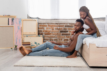 Fototapeta premium Seductive african american woman in bra touching muscular boyfriend sitting on carpet