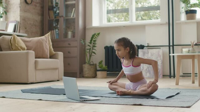 Tracking shot of cute little girl in sportswear sitting in Baddha Konasana pose on yoga mat and watching tutorial or online class on laptop