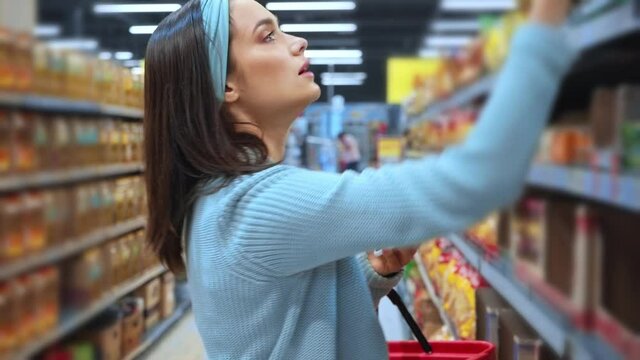 young woman holding smartphone and taking packaging from shelf in supermarket