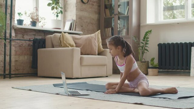 Tracking medium shot of little girl in sportswear stretching in upward facing dog pose on yoga mat at home and watching tutorial or online class on laptop