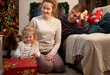 Little girl child with her young parents celebrating Christmas at home by the fireplace
