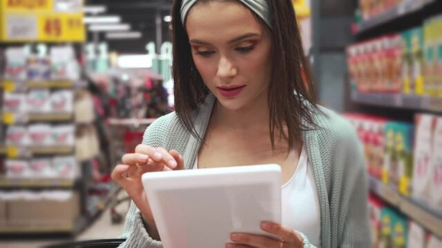 young woman checking list on digital tablet while walking in supermarket