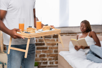 African american man holding tray with breakfast near girlfriend with book on blurred background