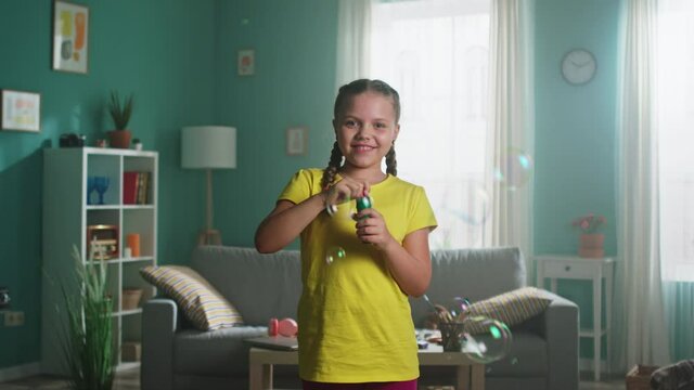 Portrait of small girl with two braids, in yellow T-shirt, smiling at camera and blowing bubbles, open-minded kid that has happy childhood, Foreground, Slow motion.