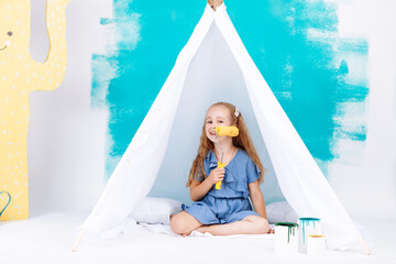 Portrait of a beautiful smiling girl with a paint roller. Little girl playing in a white wigwam on a background of colorful walls. Cheerful cute baby having fun in the nursery. baby's day. © Andriy Medvediuk
