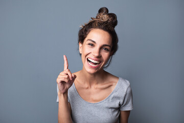 Beautiful joyful woman with a natural open smile pointing with index finger upwards over gray...