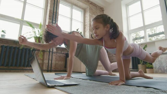 Medium shot of young mother and little daughter in sportswear kneeling on yoga mat and doing bird-dog crunches while watching online class on laptop, then resting in utthita balasana