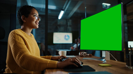 Young Multiethnic Specialist Working on Desktop Computer with Green Screen Mock Up Display in Creative Office. Beautiful Diverse Female Manager with Short Hair and Glasses is Wearing a Yellow Jumper.