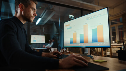 Young Handsome Project Manager Working on a Marketing Presentation on Desktop Computer in Busy Creative Office. Male Specialist is Implemeting Business Strategy Points Into the Keynote.