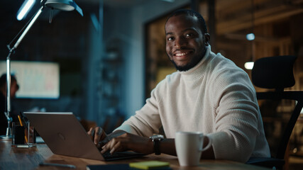 Handsome Black African American Man Having an Online Conversation on a Laptop Computer in Creative Office Environment. Happy Person of Color in Warm White Sweater is Looking at Camera and Smiling.