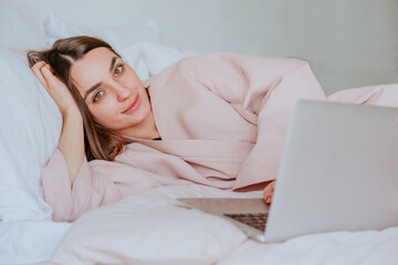 Young woman in bath robe lying on the bed using laptop for work or messaging. Morning blogger routine concept.