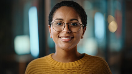 Close Up Portrait of a Young Latina with Short Dark Hair and Glasses Posing for Camera in Creative Office. Beautiful Diverse Multiethnic Hispanic Female Wearing Yellow Jumper is Happy and Smiling.