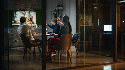 Creative Team is Having a Conference Video Call in Meeting Room Behind Glass Walls in an Office. Colleagues Sit Behind Table and Discuss Business Opportunities with Colleague on Big TV Screen.