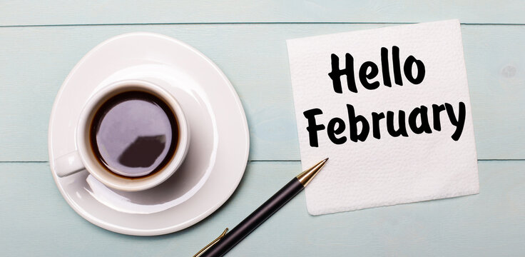 On a light blue wooden tray, there is a white cup of coffee, a handle and a napkin that says HELLO FEBRUARY