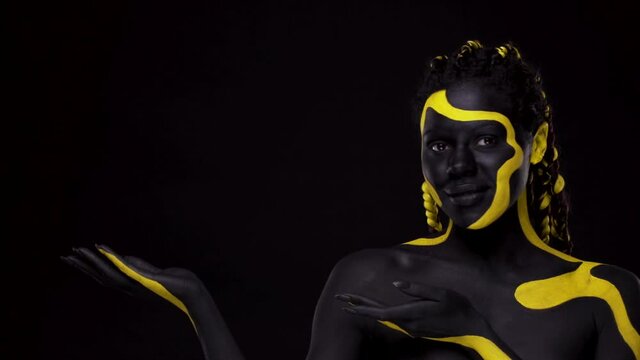 Face art. Girl showing on copy space with fingers. Woman with black and yellow body paint. Young african girl with colorful bodypaint. An amazing afro american model with makeup.