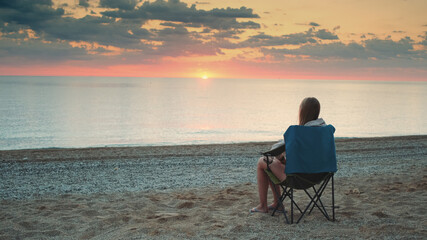 Fototapeta premium Woman admiring sunset on the sea sitting in folding tourist chair. Relaxing and enjoying nature.