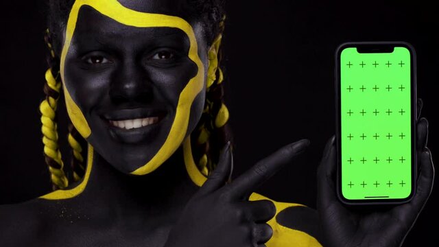Face art. Woman with black and yellow body paint. Girl pointing finger at mobile phone screen with chroma key. Young african female with colorful bodypaint. An amazing afro american model with makeup.