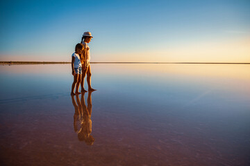 Fototapeta premium Two little lovely happy sisters are walking along the mirror-like pink salt lake enjoying the warm summer sun in the long-awaited vacation